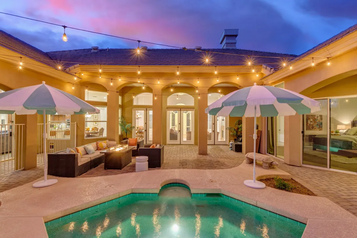 This vibrant exterior image features a swimming pool surrounded by comfortable outdoor seating, umbrellas, and string lights. The setting suggests both daytime fun and evening relaxation, allowing guests to envision themselves enjoying the backyard across different seasons. The seating area and fire pit in the background highlight the space's versatility for alfresco meals and cool nights outdoors.