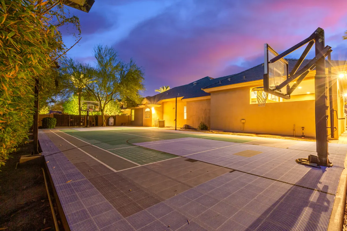 A well-maintained basketball court, complete with hoop and surrounded by mature trees and evening lighting, invites energetic play and family sports matches in the backyard.