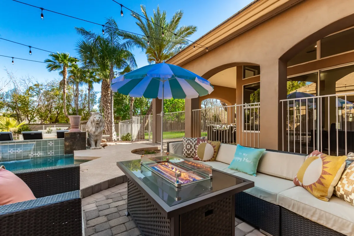 A cozy fire pit area with ample lounge seating sits adjacent to a shimmering pool, surrounded by palm trees and playful details like a colorful umbrella and lion statue. The backdrop offers a welcoming space for group gatherings, dining, and relaxing by the fire under the stars.