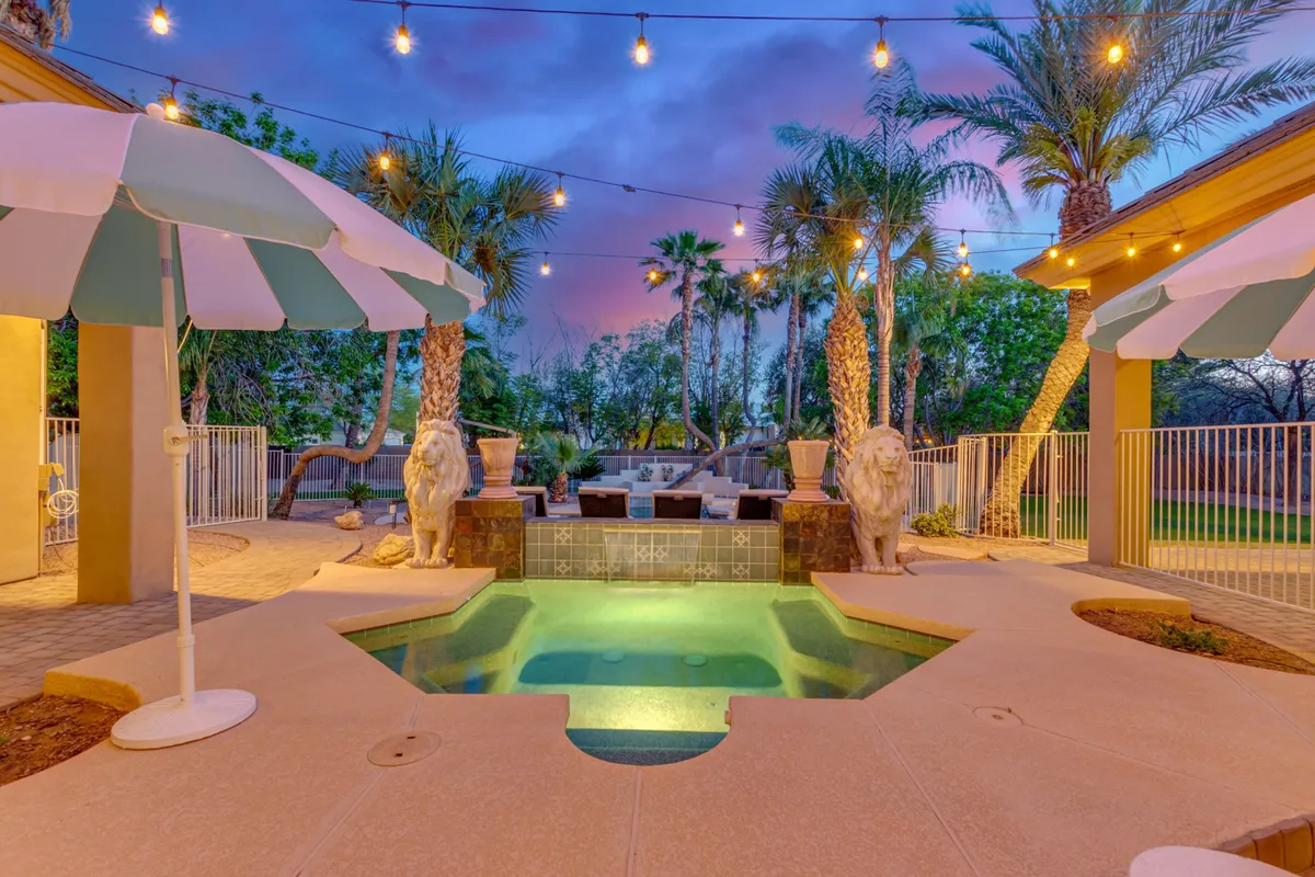 A sparkling swimming pool forms the focal point, bordered by elegant decorative lion statues and surrounded by lush palm trees and outdoor lounge furniture. String lights set a festive, magical mood as the sun sets, with the entire area designed for evening gatherings and relaxation.