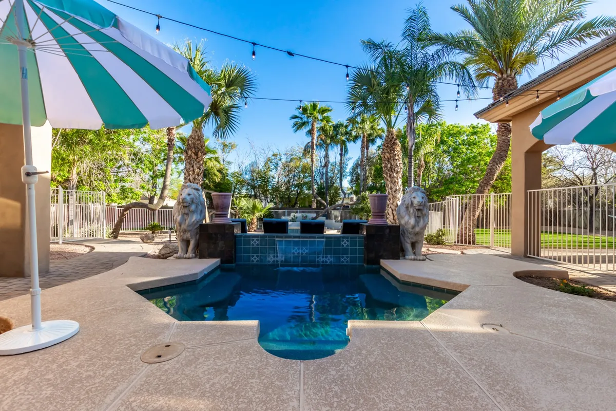 A serene pool scene with classic lion statues, palm trees, and umbrellas, creating a luxurious and tranquil environment. The seating area nearby is perfect for relaxing or entertaining, set against lush landscaping.