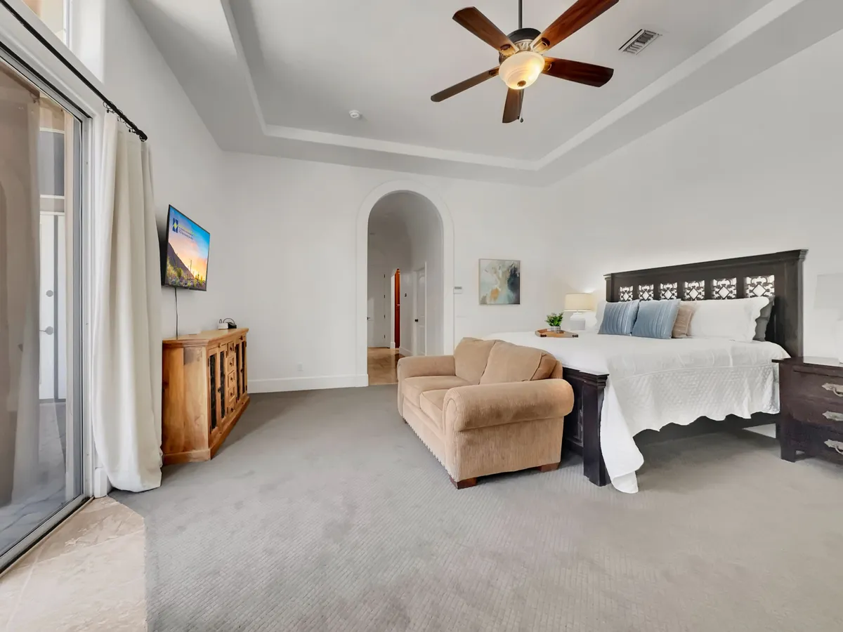 A serene bedroom featuring a plush bed, a comfortable sofa, a wall-mounted TV, a wooden dresser, and an arched doorway. Soft lighting and neutral tones create a cozy, restful atmosphere ideal for relaxing mornings or unwinding after a long day.