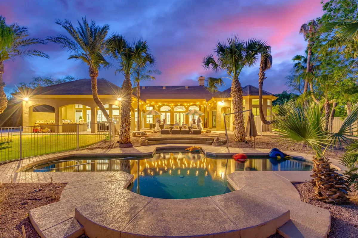 The image showcases the stunning backyard of a luxury single-family home in Chandler, Arizona. The focal points include a sparkling swimming pool surrounded by lush palm trees, a stylish outdoor seating area perfect for gatherings or relaxation, and the home's beautifully illuminated exterior that creates a warm, inviting glow as the sun sets. The scene exudes tranquility, offering glimpses of serene poolside evenings and memorable outdoor dining experiences.