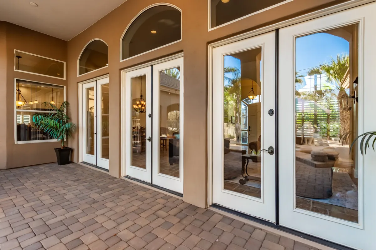 The home's elegant double doors, arched windows, and a charming brick pathway set an inviting scene for guests. Lush potted plants and shaded areas promise a warm welcome and hint at the hospitality within.